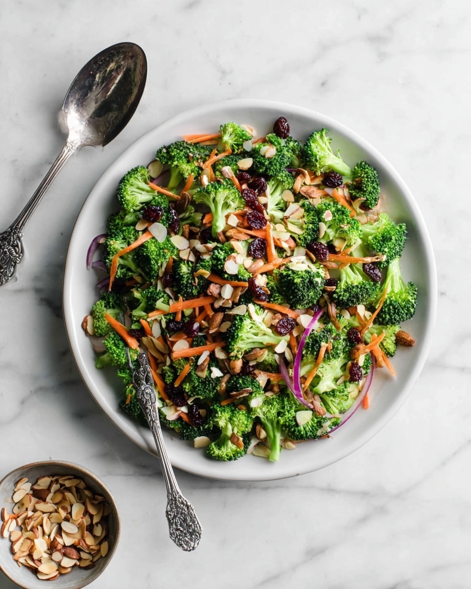 A white plate filled with a fresh broccoli salad sits on a white marbled surface. The salad has bright green broccoli florets as the main layer, mixed with thin orange carrot strips and small pieces of red onion scattered throughout. There are dark dried cranberries and thin almond slivers spread evenly on top, adding texture and color contrast. A detailed silver spoon rests on the left side of the plate, and a small bowl with extra almond slivers is placed below the plate. The photo taken with an iphone --ar 4:5 --v 7