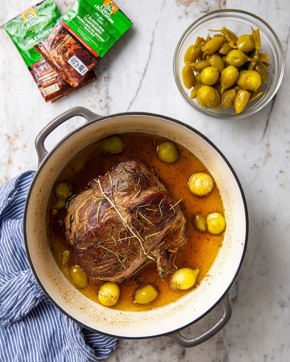 A round white cooking pot with dark edges contains one large, brown roasted meat piece tied with string in the center, sitting in a pool of golden-brown sauce; around the meat, several small yellow-green peppers float in the sauce. Nearby, there is a clear bowl filled with similar yellow-green peppers, and in the background on a white marbled surface, two green and brown seasoning packets are partially visible. A blue and white striped cloth is placed on the lower left corner. Photo taken with an iphone --ar 4:5 --v 7