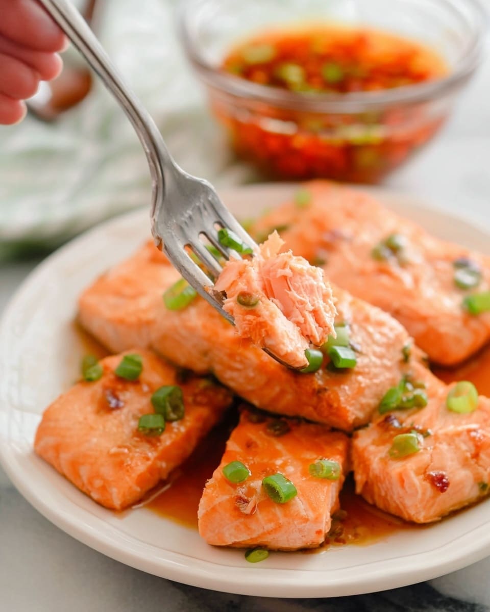 The image shows a white plate filled with several pieces of cooked salmon, each piece light orange with a slightly glazed texture and scattered green onion slices on top. A woman's hand is holding a fork that lifts a small piece of salmon, showing its soft, flaky inside. In the blurred background, there is a clear glass bowl with an orange-red sauce containing small green pieces. The whole scene is set on a white marbled surface. photo taken with an iphone --ar 4:5 --v 7