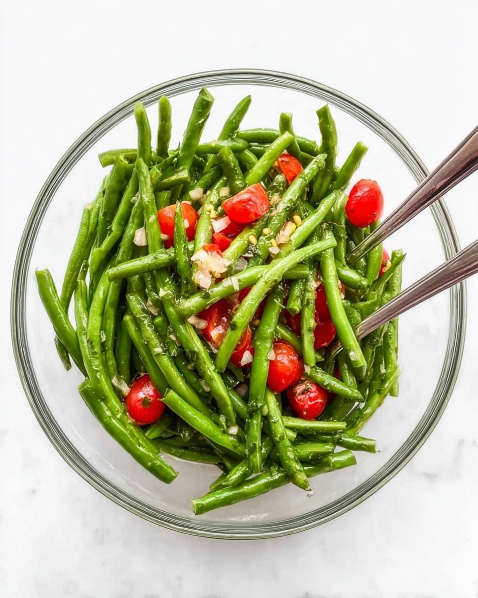 A clear glass bowl filled with bright green beans that are whole and neatly laid out, mixed with small pieces of red cherry tomatoes and finely chopped light-colored onions scattered on top and throughout the beans, with two metal spoons placed inside the bowl on opposite sides for serving, all set on a white marbled surface photo taken with an iphone --ar 4:5 --v 7