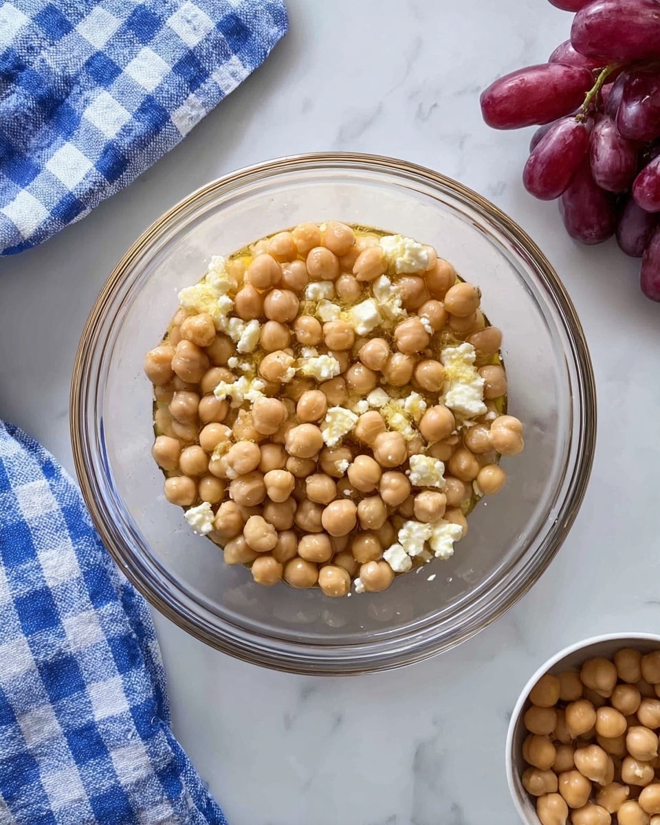 A clear glass bowl sits on a white marbled surface, filled with a mix of light brown chickpeas and small white cheese chunks coated lightly in oil. To the right, there is a small white bowl filled with more chickpeas and bunches of red grapes. On the left side, a blue and white checkered cloth is partially visible. The overall image is bright and clean. photo taken with an iphone --ar 4:5 --v 7