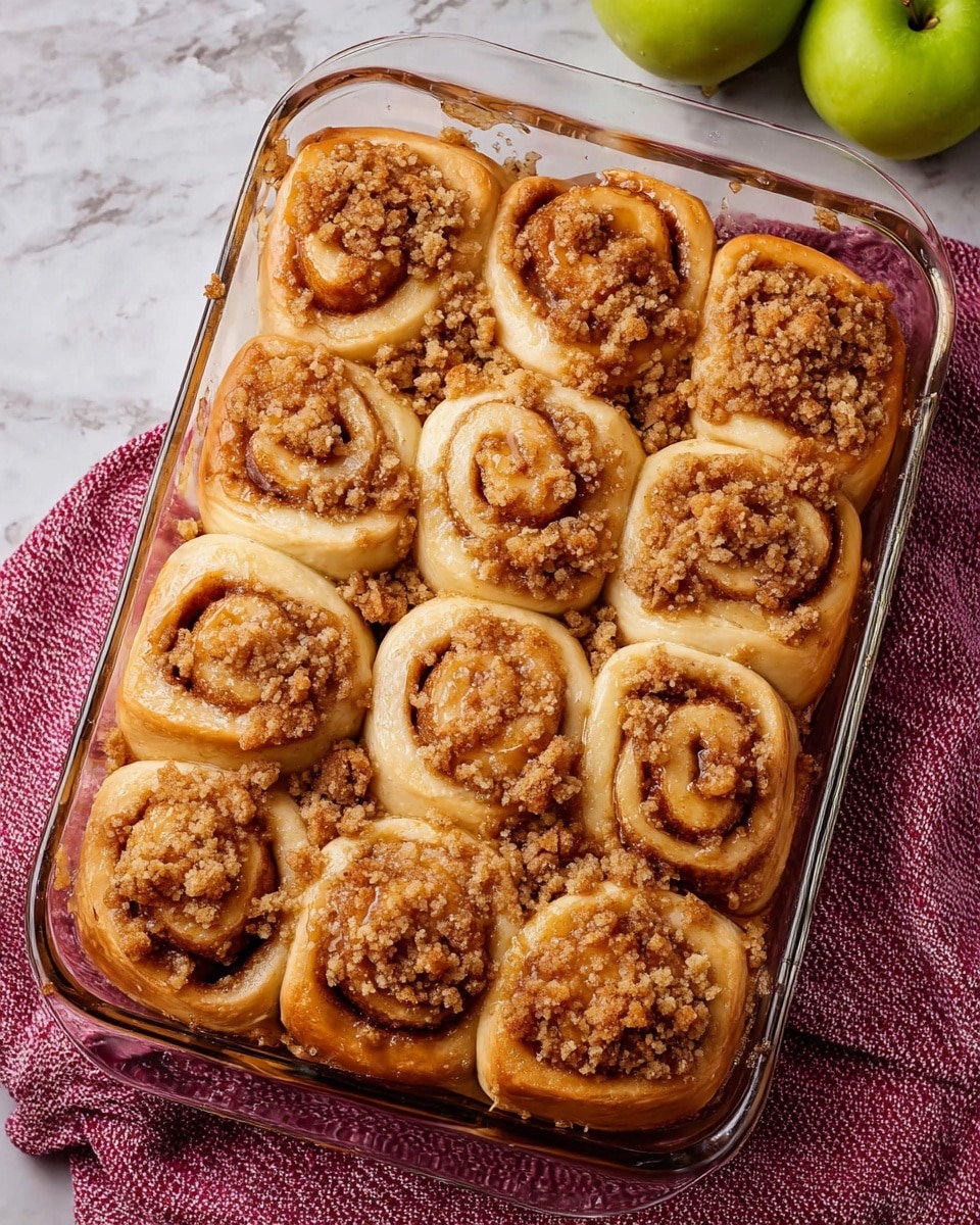 A clear glass rectangular baking dish filled with twelve golden brown cinnamon rolls, arranged in three rows of four. Each roll has a swirled shape with a light brown cinnamon filling visible in the layers, topped generously with crumbly streusel pieces. The rolls have a soft and fluffy texture with a slightly shiny, baked surface. The dish is set on a white marbled surface with a pink textured cloth underneath, and a few green apples are partially visible in the background. photo taken with an iphone --ar 4:5 --v 7