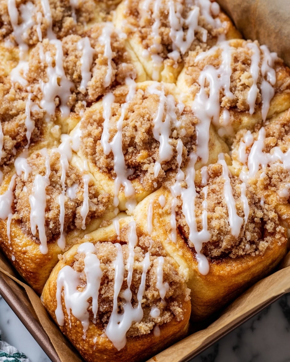 The image shows a close-up of a tray filled with soft, golden-brown rolls arranged tightly next to each other. Each roll is topped with a crumbly, light brown streusel layer that looks crunchy and textured. Over this, white icing is drizzled unevenly, creating thin lines that run in different directions. The tray is lined with brown parchment paper, and the background surface is a white marbled texture. photo taken with an iphone --ar 4:5 --v 7