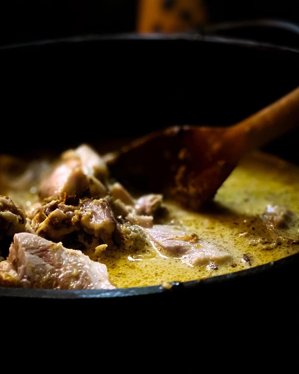 The image shows a close-up of a pan with a dark rim filled with a creamy, yellowish liquid, possibly a sauce or broth. Large pieces of white and light brown meat are visible floating in the liquid, with some texture detail on the meat. A wooden spoon is stirring or lifting some of the meat from the liquid. The scene is dimly lit, focusing on the food in the pan, with a blurred background. Photo taken with an iphone --ar 4:5 --v 7