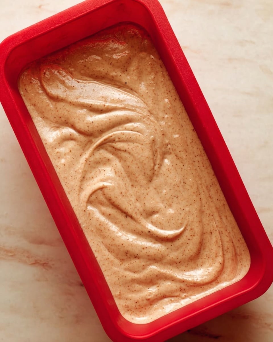 This image shows a red rectangular silicone mold filled with a creamy light brown batter that has small darker brown specks throughout. The batter is smooth with soft swirls on the surface, giving it a slightly textured look. The mold is resting on a white marbled surface. photo taken with an iphone --ar 4:5 --v 7