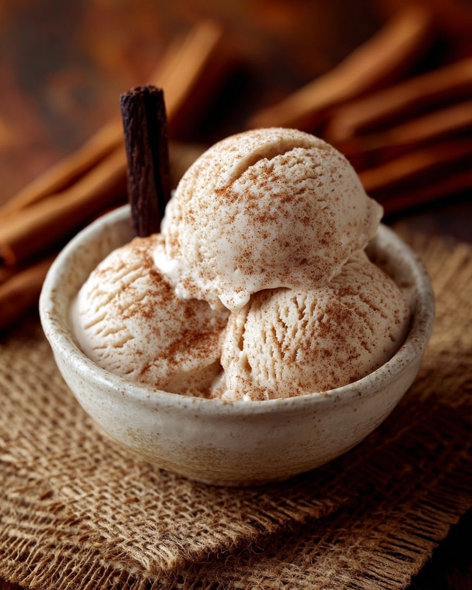 A scoop of light brown ice cream with visible cinnamon specks is placed inside a small white bowl. The ice cream has a creamy texture with subtle ridges and soft peaks. Two cinnamon sticks lean against the side of the bowl, with additional cinnamon sticks scattered around the bowl on a white marbled surface partly covered by a textured beige woven fabric and a red checkered cloth. The warm tones of the cinnamon and fabric contrast with the bowl and the ice cream. Photo taken with an iphone --ar 4:5 --v 7