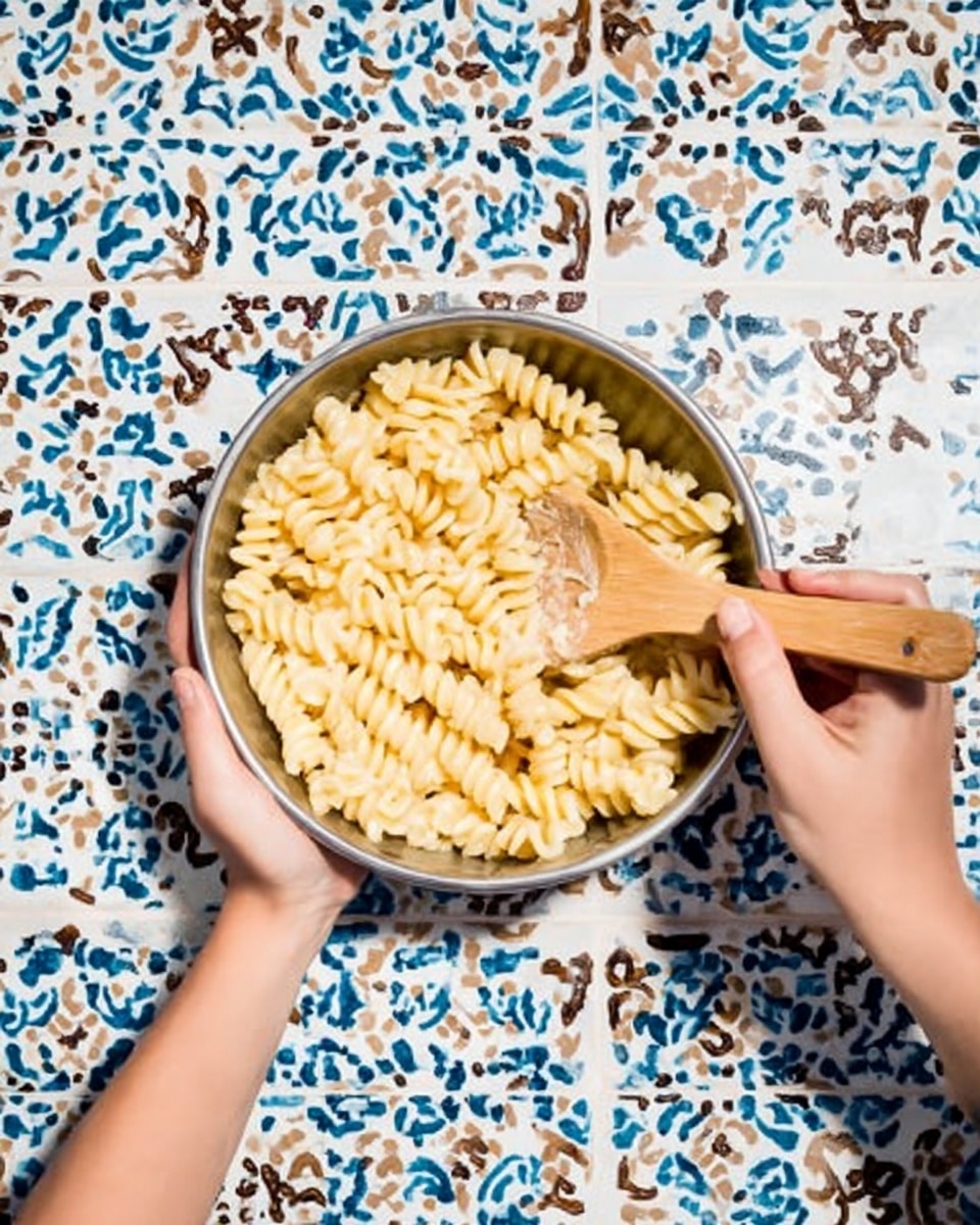 A metal bowl filled with creamy spiral pasta sits in the center of a white marbled surface, held by a woman's hand on each side. A wooden spoon stirs the pasta, which is light yellow with a smooth texture and coated in sauce. The background shows colorful blue, white, and brown patterned tiles. photo taken with an iphone --ar 4:5 --v 7