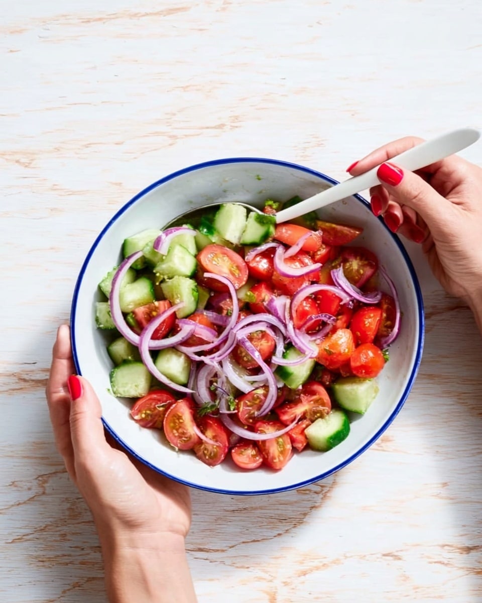 A bright salad is shown inside a white bowl with a blue rim, placed on a white marbled surface. The salad has three main layers: at the bottom, there are fresh green cucumber cubes with smooth skin; the middle layer is made of shiny red tomato pieces scattered evenly; and on top, thin purple onion slices curl in loose rings. A woman's right hand holds a white spoon on the left side of the bowl, and her left hand steady the bowl from the right, both hands with red nail polish. The scene is well lit, highlighting the natural colors of the vegetables. photo taken with an iphone --ar 4:5 --v 7
