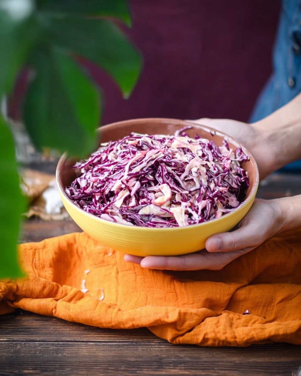 A yellow bowl filled with a large pile of creamy shredded cabbage salad showing layers of purple and pale pink cabbage mixed together. The bowl is held by woman's hands resting on a bright orange cloth, placed on a brown wooden surface. On the left side, there is a green plant with large leaves, and the background is a simple dark purple. The scene captures a close-up view with natural light highlighting the creamy texture of the salad photo taken with an iphone --ar 4:5 --v 7