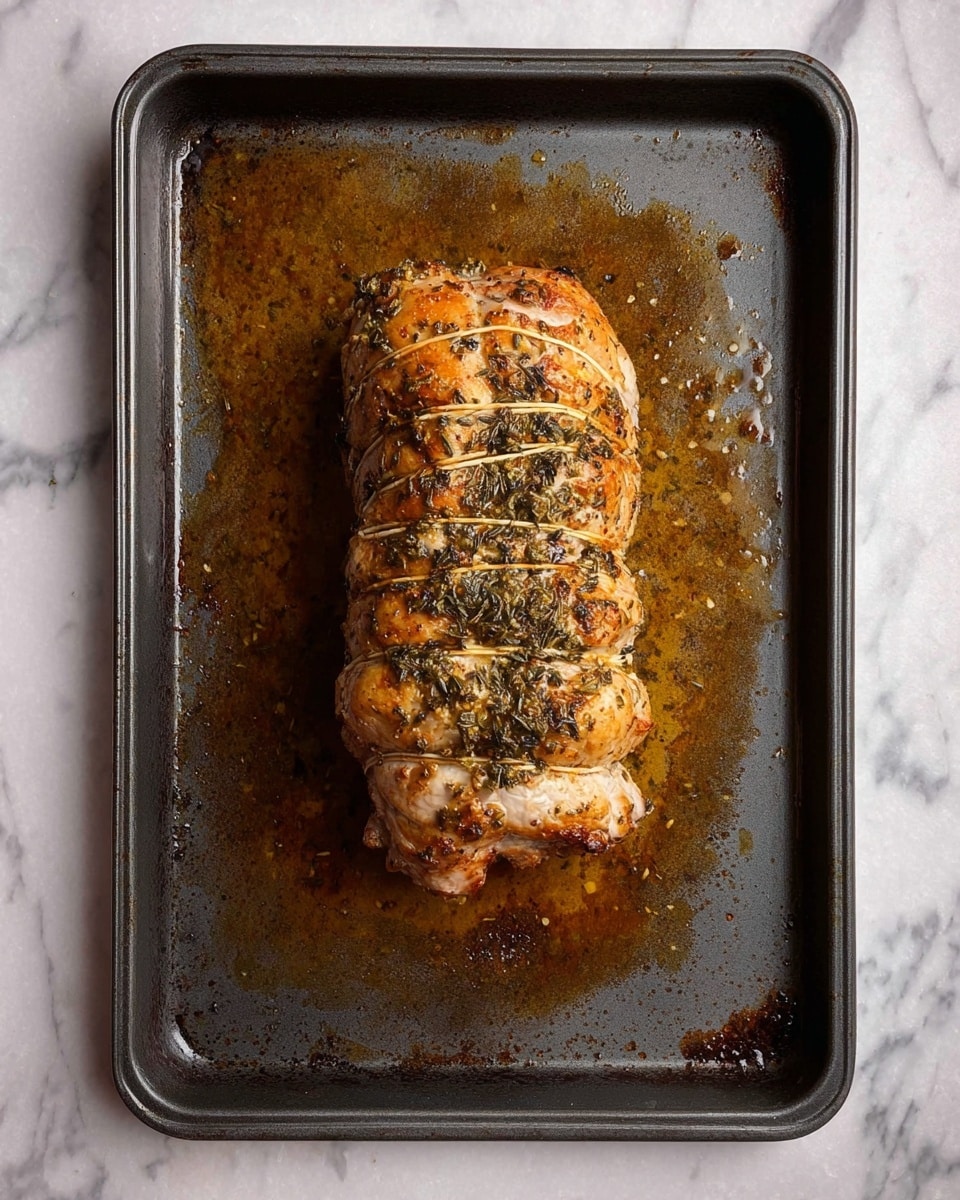 A single roasted meat roll tied with string, sitting in the middle of a dark metal baking tray with browned oil and seasoning residues spread around it; the meat has a golden brown color with visible herb and spice coating on top, showing a rough textured surface and slight crisping on the edges; the background is a white marbled surface. photo taken with an iphone --ar 4:5 --v 7