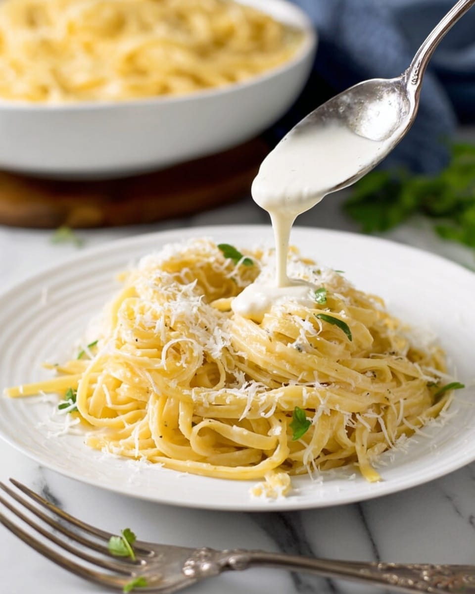 A white plate filled with a nest of creamy yellow pasta strands piled high, lightly sprinkled with white grated cheese and small green herb leaves, while a spoon above pours smooth white sauce dripping over the pasta, all sitting on a white marbled surface, with a blurred bowl of more pasta in the background and a silver fork placed beside the plate, photo taken with an iphone --ar 4:5 --v 7