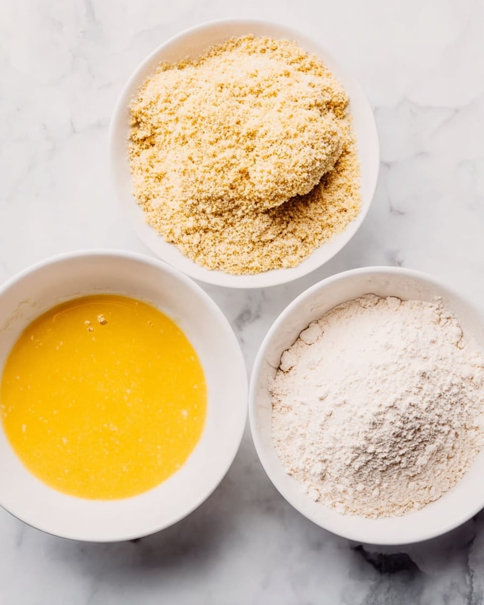 The image shows three white bowls arranged on a white marbled surface. The top bowl contains pieces of food covered in light golden breadcrumbs, creating a rough texture. The left bowl is filled with a smooth, bright yellow egg mixture that looks glossy. The bowl on the right contains fine, white flour with a powdery texture, spread evenly. The bowls are placed close together, showing a clear process of coating food items in flour, then egg, and finally breadcrumbs. photo taken with an iphone --ar 4:5 --v 7
