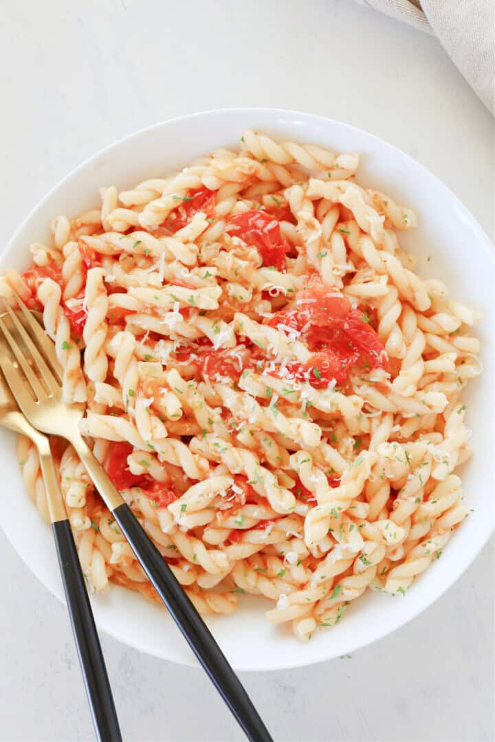 A white bowl filled with twisted pasta, creamy light orange in color, mixed with bright red tomato pieces scattered throughout. The pasta has finely chopped green herbs sprinkled on top and a light dusting of grated white cheese. Two gold forks with black handles rest on the left side inside the bowl. The bowl is placed on a white marbled surface. Photo taken with an iphone --ar 4:5 --v 7