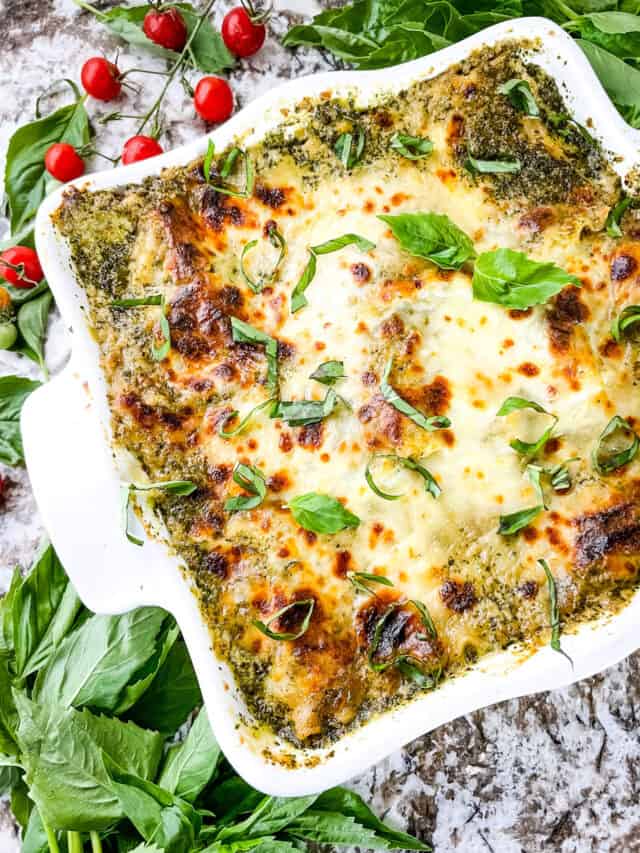 Lasagna al Pesto Recipe 4 A square white baking dish contains a baked dish with two visible layers: a green pesto layer spread evenly underneath and topped with melted mozzarella cheese that is golden brown with bubbly spots and some toasted edges. Fresh green basil leaves are scattered over the top, adding a bright contrast. The dish sits on a white marbled surface with fresh basil leaves and small red cherry tomatoes placed decoratively around it. Photo taken with an iphone --ar 4:5 --v 7