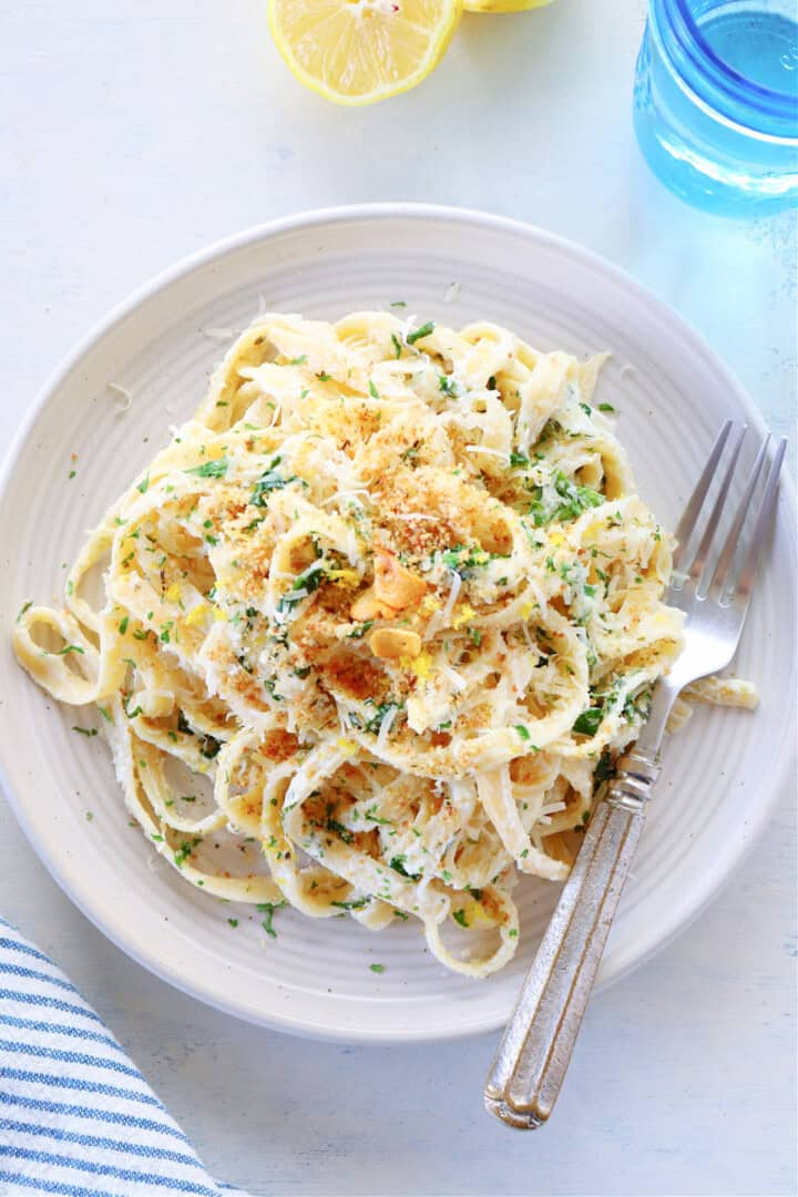 A white plate with creamy fettuccine pasta arranged in a loose mound as the main layer, coated in a smooth white sauce. On top, there is a light sprinkling of golden brown toasted breadcrumbs and green chopped herbs. Thin slices of garlic are scattered among the pasta strands, adding texture. A silver fork rests on the right side of the plate. The plate is placed on a white marbled surface, with a lemon half visible in the top background and a blue glass of water to the right. The bottom left corner shows a blue and white striped cloth. Photo taken with an iphone --ar 4:5 --v 7