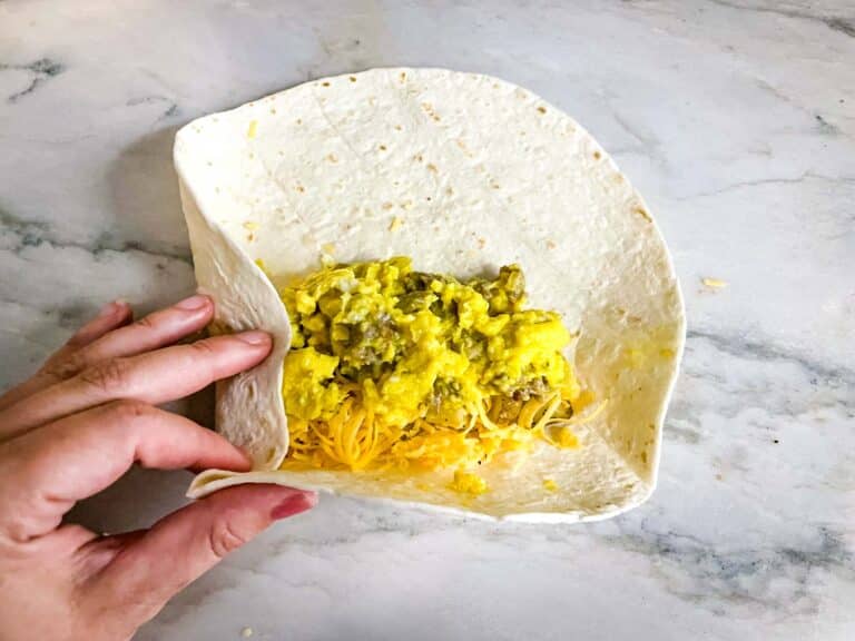 A white flour tortilla is opened and held by a woman's hand on the left side, revealing two layers inside; the bottom layer consists of shredded yellow cheese, and the top layer is scrambled eggs mixed with browned sausage pieces, all placed on a white marbled surface. photo taken with an iphone --ar 4:5 --v 7