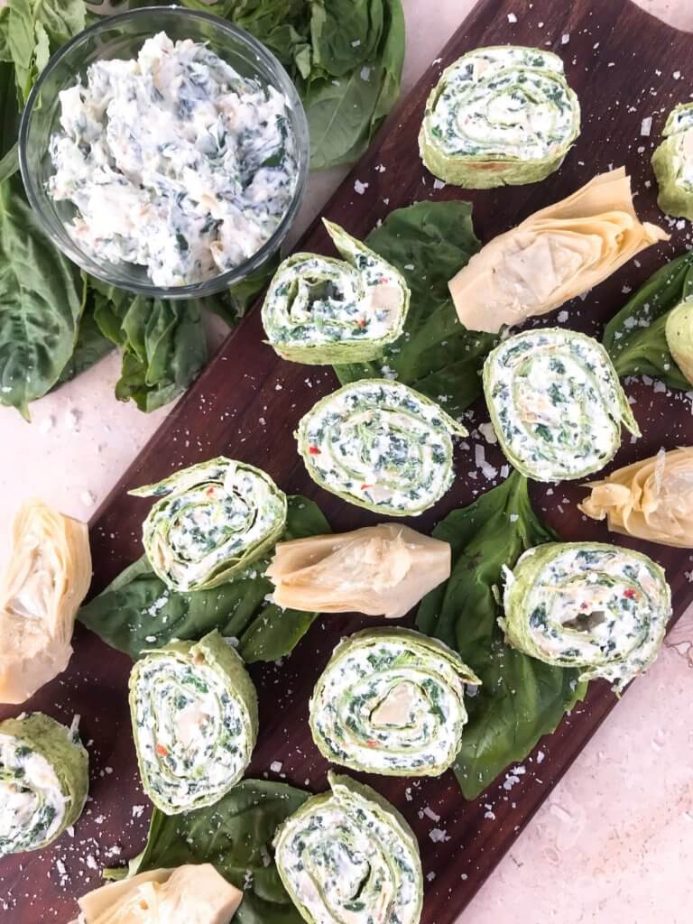 A wooden board is laid on a white marbled surface, topped with green spinach leaves as a base. On these leaves, there are several small, round rolled pinwheels showing three visible layers: a light green outer layer, a white creamy middle layer mixed with green herbs, and visible bits of red seasoning inside. Scattered among the pinwheels are light beige artichoke pieces with a soft texture. In the top left corner, there is a clear glass bowl filled with a creamy white mixture containing green herbs. Coarse salt is sprinkled lightly over the spinach leaves and pinwheels. The setting is bright and clean with soft natural light. Photo taken with an iphone --ar 4:5 --v 7