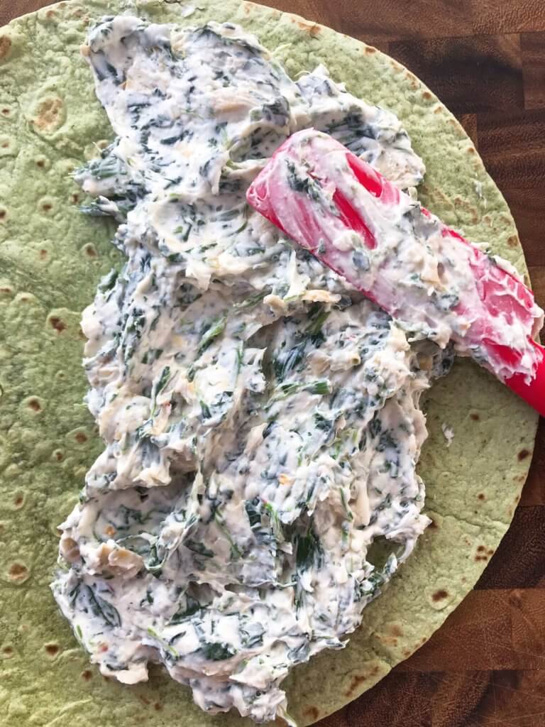 A green spinach wrap is placed flat on a white marbled surface, topped with a thick layer of creamy white mixture that has visible green leafy bits and small chunks throughout. A red silicone spatula smeared with the same creamy mixture rests on top of the spread, showing the texture as soft and slightly chunky. The wrap and mixture fill most of the image, highlighting the contrast between the green wrap and the white mixture. Photo taken with an iphone --ar 4:5 --v 7