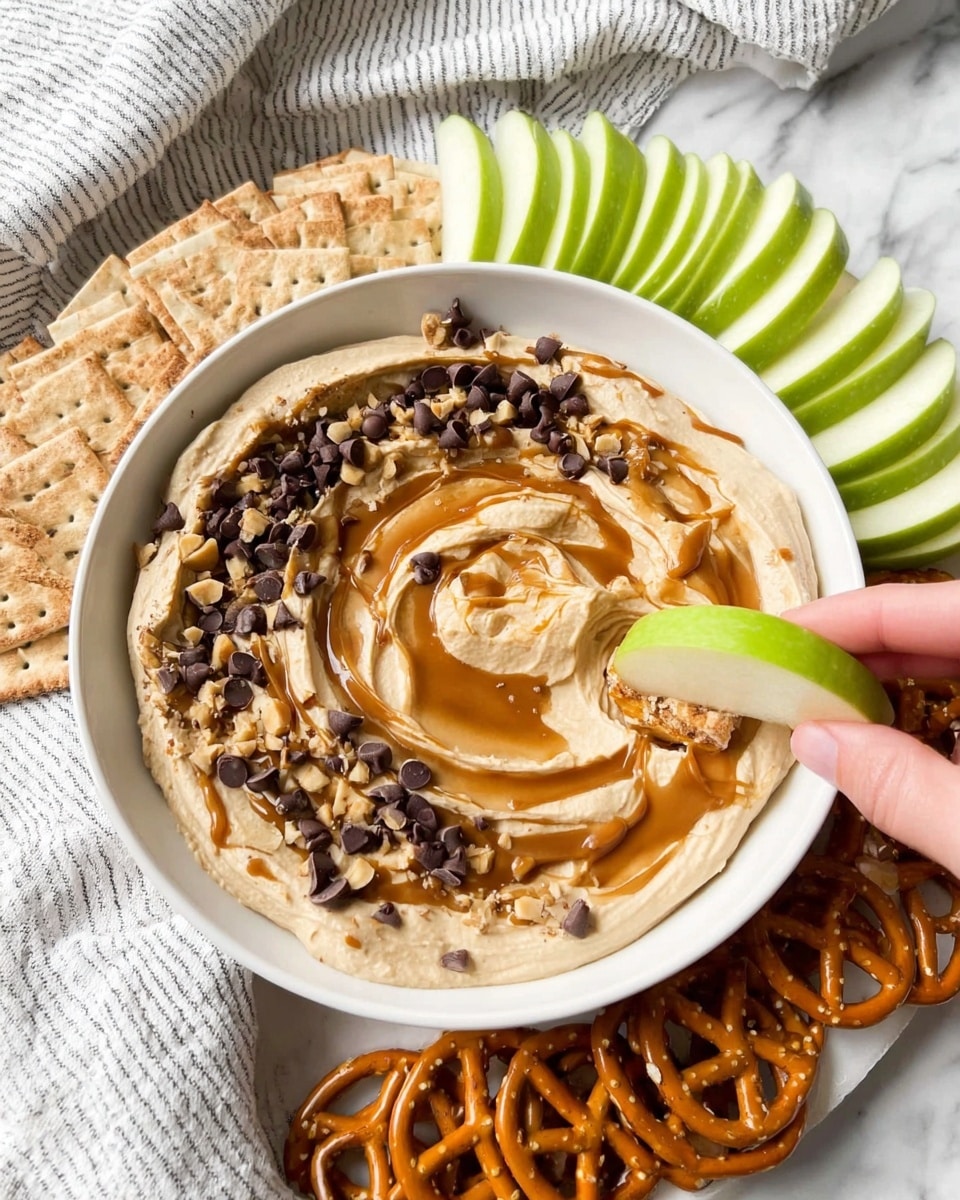 A white bowl filled with a creamy light brown dip as the bottom layer, topped with a swirl of caramel sauce, scattered small dark chocolate chips, and chopped peanuts. Around the bowl, there are neatly arranged green apple slices, rectangular light brown crackers, and small pretzels forming three separate groups on a white marbled surface. A woman's hand is holding one green apple slice dipped with some of the creamy dip, caramel, chocolate chips, and peanuts. The setting includes a white and gray striped cloth in the background. Photo taken with an iphone --ar 4:5 --v 7