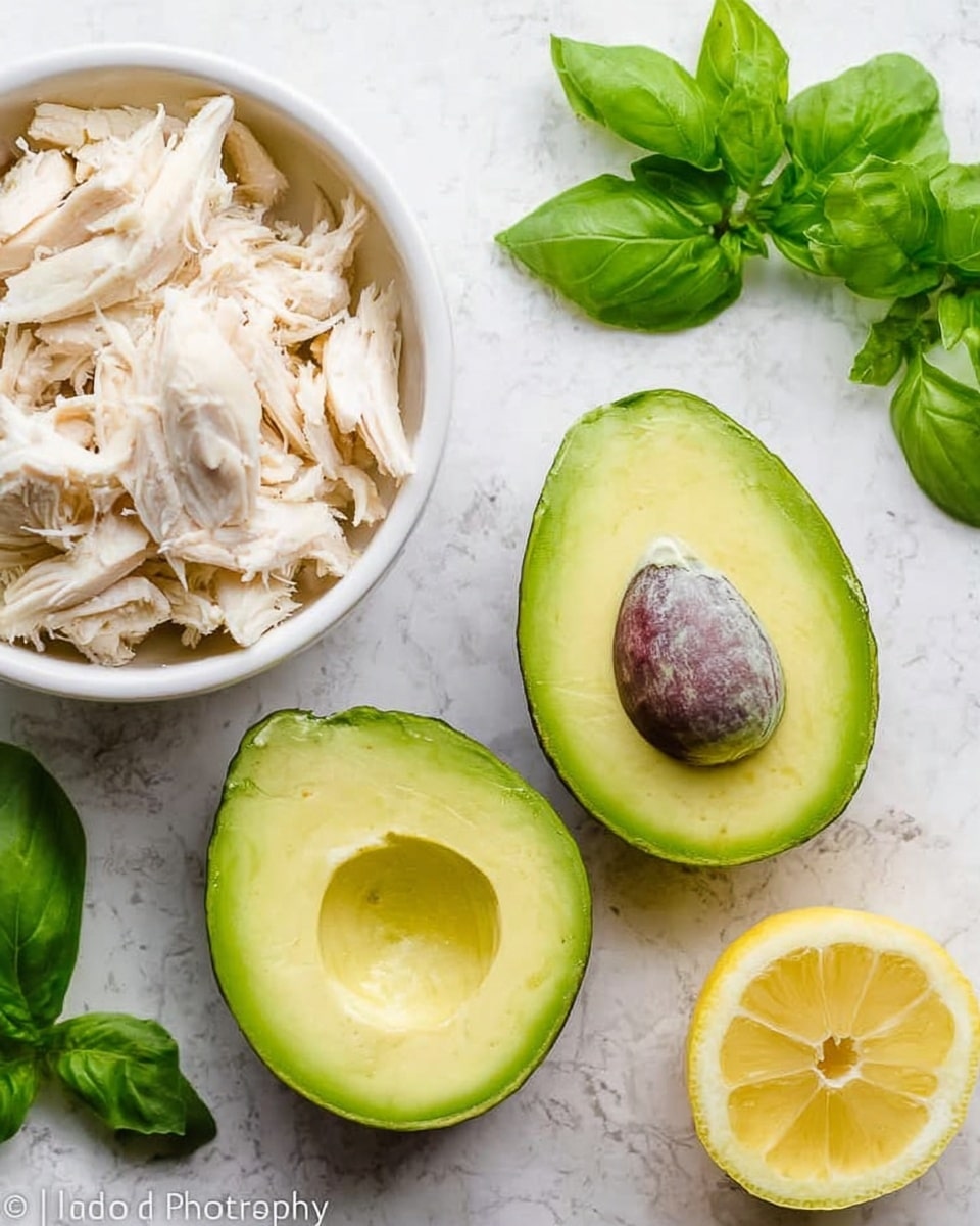The image shows two halves of a ripe avocado with dark green skin and bright yellow-green flesh, placed on a white marbled surface. Above the avocado halves, there is a white bowl filled with shredded white chicken meat. To the left and right of the avocado, fresh green basil leaves are scattered around. On the right, a half lemon with bright yellow flesh and a textured rind sits on the surface. The whole setup is bright and clean, focused on fresh ingredients. photo taken with an iphone --ar 4:5 --v 7