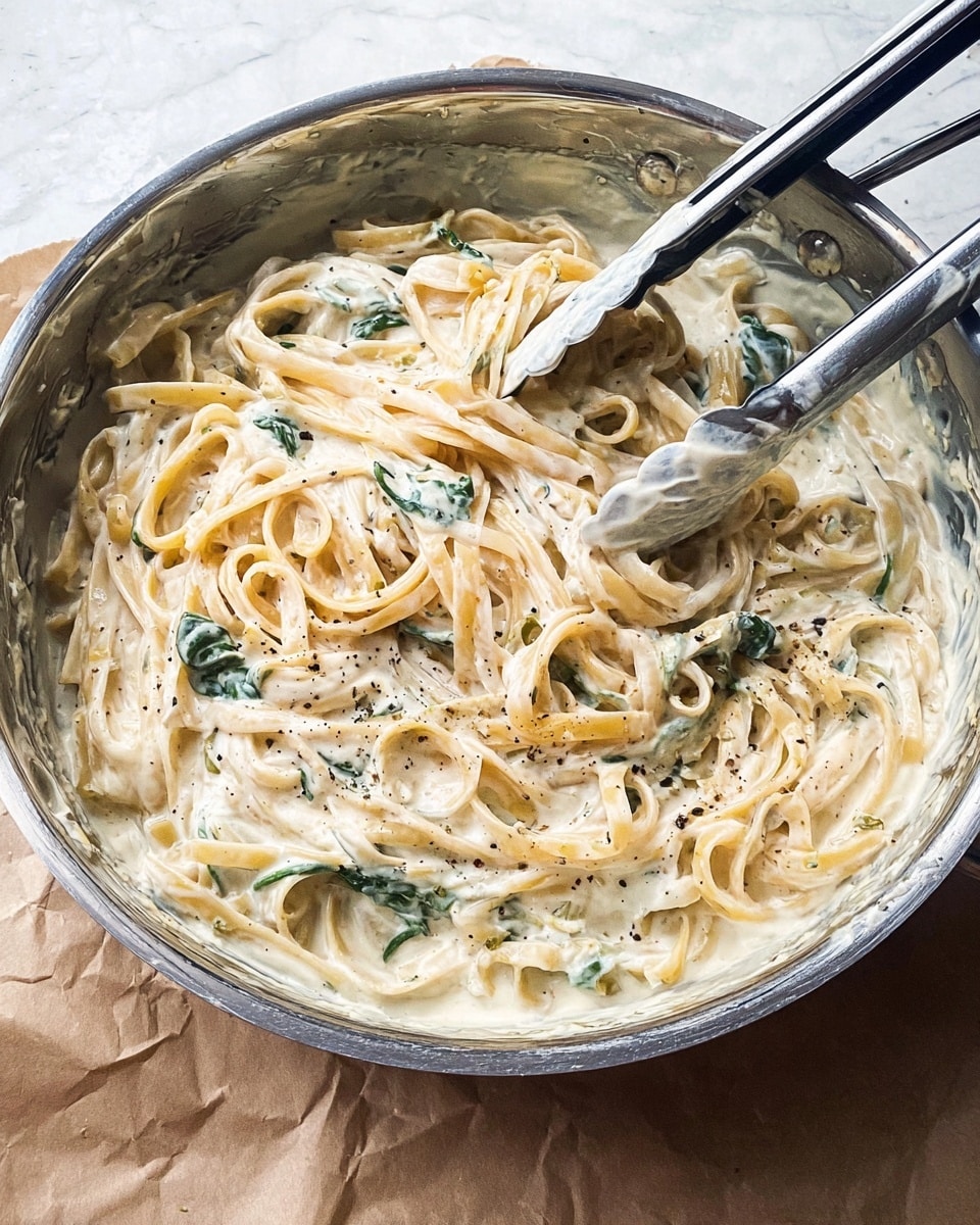 A close-up view of a metal pan filled with creamy pasta, showing two silicone-tipped metal tongs resting inside. The pasta is long and flat, coated evenly in a thick white sauce with visible flecks of black pepper. There are some green leafy pieces mixed throughout the pasta, adding color contrast. The pan sits on a light brown, crumpled paper surface, but the background has been changed to a white marbled texture. The scene looks simple and focused on the textured creamy pasta and sauce. Photo taken with an iphone --ar 4:5 --v 7