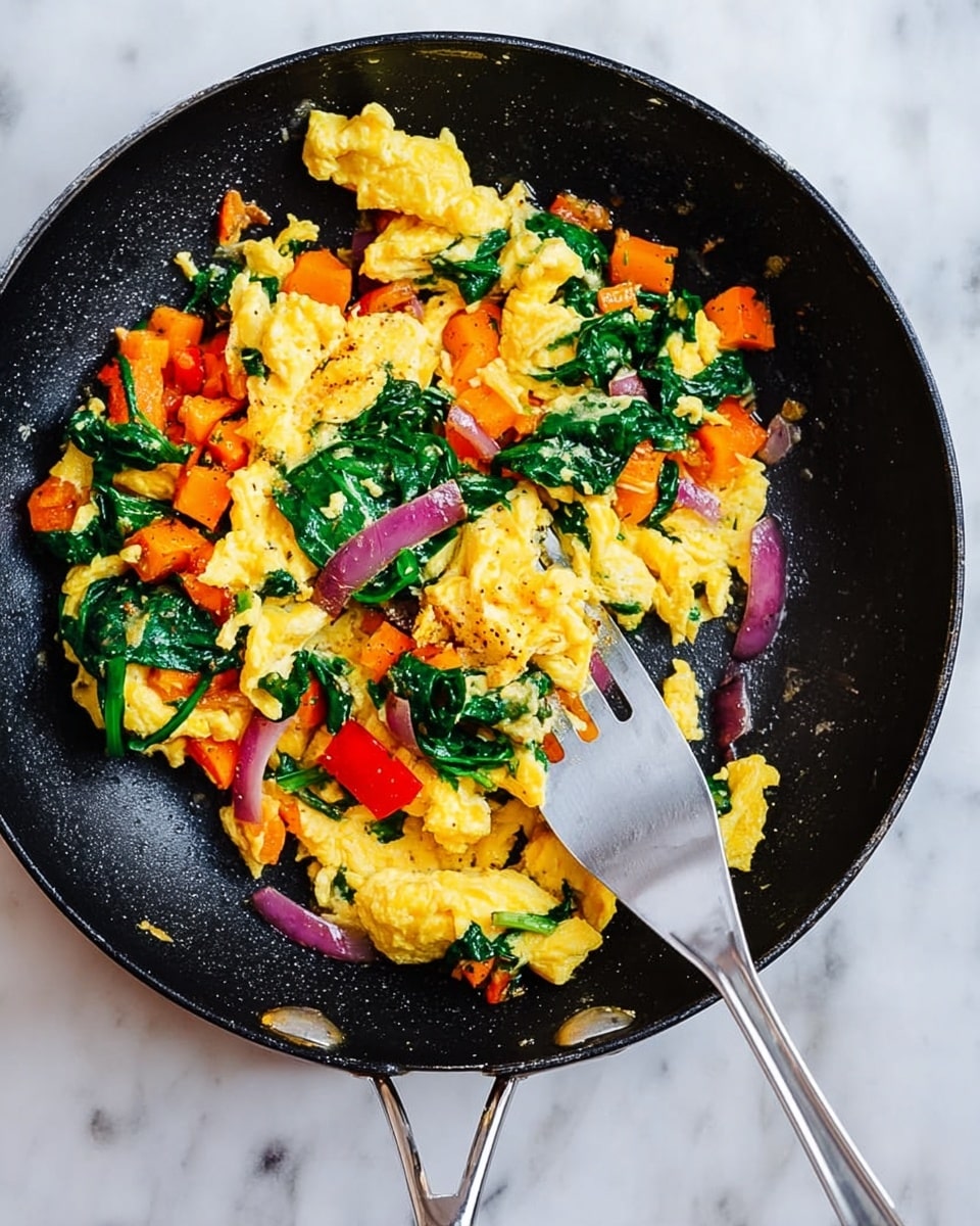 In a black pan on a white marbled surface, there is a mix of scrambled yellow eggs with soft texture, bright green leafy spinach, orange chunks of carrot, and pieces of red bell pepper, all stirred together. There are also some thin slices of purple onion spread around. A silver spatula rests diagonally inside the pan, catching some of the mix. The overall look is colorful with a nice balance of yellow, green, orange, red, and purple photo taken with an iphone --ar 4:5 --v 7
