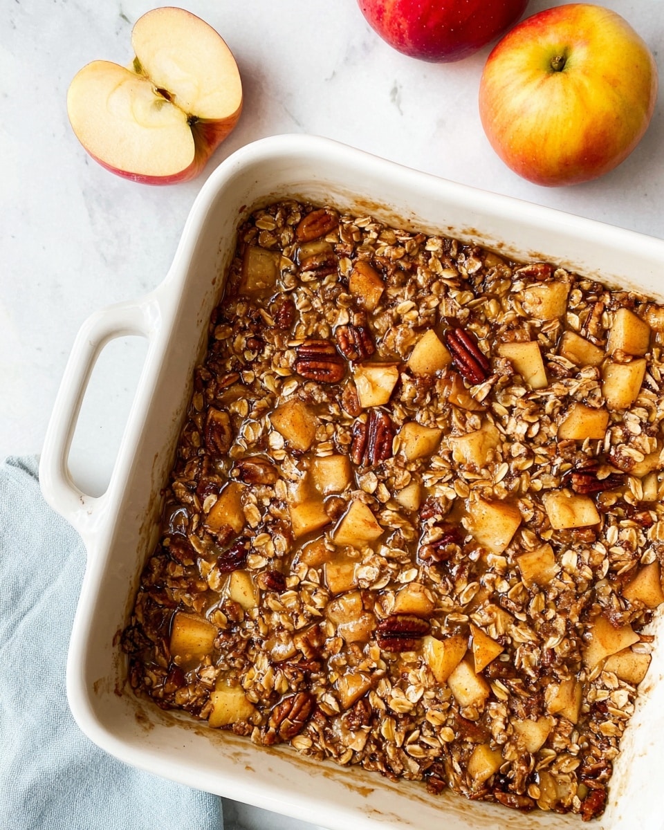 A white square baking dish filled with one thick layer of mixed oatmeal and small diced apples in a warm golden-brown syrup, with visible toasted pecans scattered throughout. The top is textured with oats and apple pieces, some showing reddish skin while others are a light yellow. The dish sits on a white marbled background with two red-yellow apples and a light blue cloth partially visible on the left side. The edges of the baking dish have slight browning from baking photo taken with an iphone --ar 4:5 --v 7