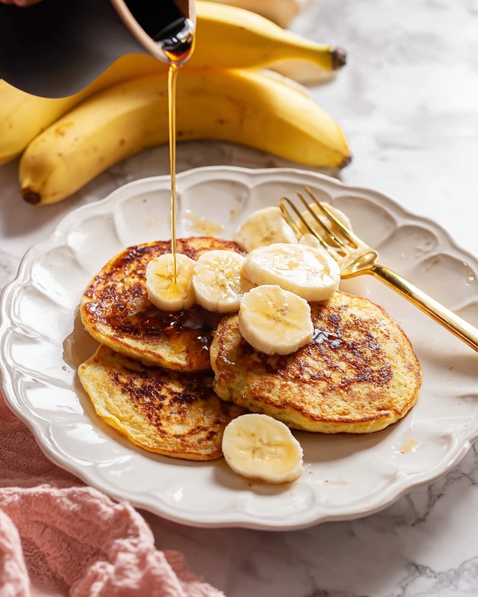 A white scalloped plate holds three golden brown pancakes arranged in a loose stack, each pancake showing a slightly uneven texture with browned spots. Sliced banana pieces are placed on top and around the pancakes, adding pale yellow and soft texture contrast. Dark syrup is being poured over the pancakes from a small, black pitcher, creating a shiny, flowing layer that pools lightly on the surface. A gold fork rests on the right side of the plate near the banana slices. The plate sits on a white marbled surface with two whole bananas and a pink cloth nearby in soft focus. Photo taken with an iphone --ar 4:5 --v 7