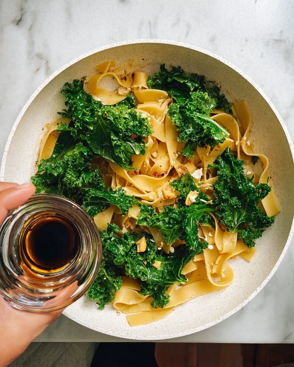 In a white speckled pan, wide, flat pale yellow pasta strips are mixed with bright green kale leaves that have a rough, leafy texture. There are small bits of light-colored ingredients scattered between the pasta and kale. In the lower left corner, a woman's hand is holding a small clear glass bowl filled with a dark brown sauce, ready to pour over the pasta and greens. The background shows a white marbled surface around the pan. photo taken with an iphone --ar 4:5 --v 7