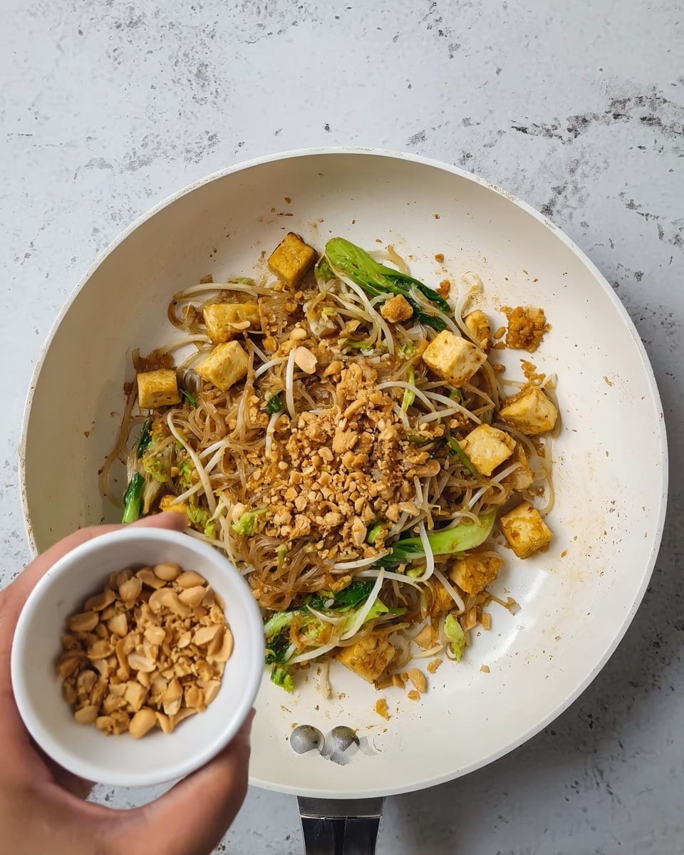 A white frying pan contains a dish with three main layers visible: the bottom layer is light brown noodles mixed with some bean sprouts, the middle layer has green leafy vegetables cut into stripes scattered around, and the top layer shows golden browned small tofu cubes spread evenly on top. A woman's hand is holding a small white cup filled with chopped light brown nuts or crushed peanuts, positioned near the pan as if ready to add them. The pan is on a white marbled surface. photo taken with an iphone --ar 4:5 --v 7