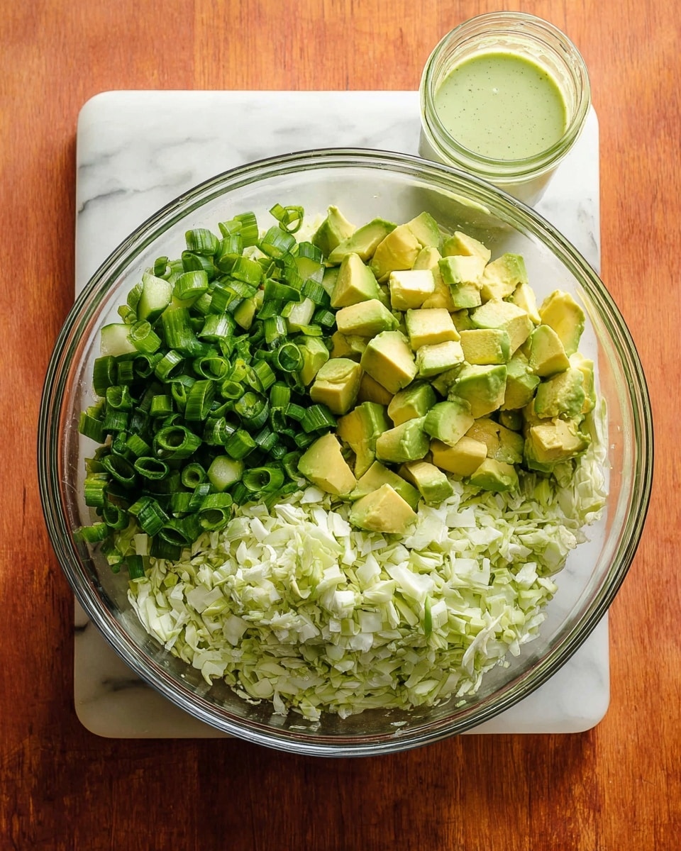 A clear glass bowl sits on a white marbled surface filled with four layers of chopped vegetables. The bottom layer has light green chopped cabbage with a rough texture. Above this is a middle layer of finely chopped pale green cucumber. To the left, there is a pile of dark green sliced spring onions with a smooth, tubular texture. On top in the center, there are chunks of light yellow-green avocado with skin on some pieces. Next to the bowl on the right side, a small clear glass jar holds a light green creamy dressing. The photo was taken with an iphone --ar 4:5 --v 7