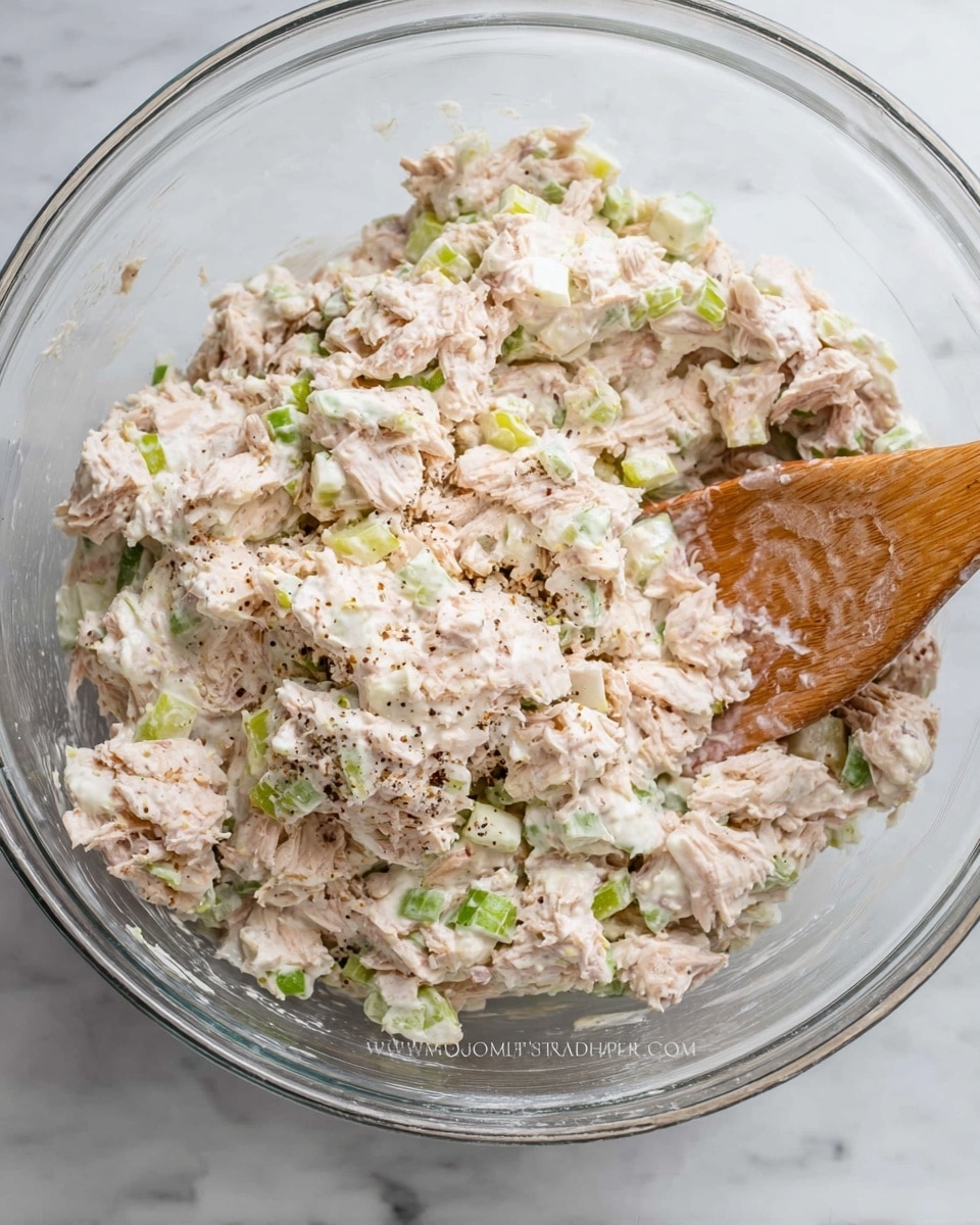 A clear glass bowl filled with a creamy tuna salad mixture, showing a mix of pale beige tuna flakes, bright white chopped onions, and light green celery pieces all coated in a thick creamy dressing. A wooden spoon rests in the bowl at the right side, partially covered by the salad, while the inside of the bowl and the salad show a somewhat chunky texture with visible bits of pepper sprinkled on top. The background features a white marbled surface. photo taken with an iphone --ar 4:5 --v 7