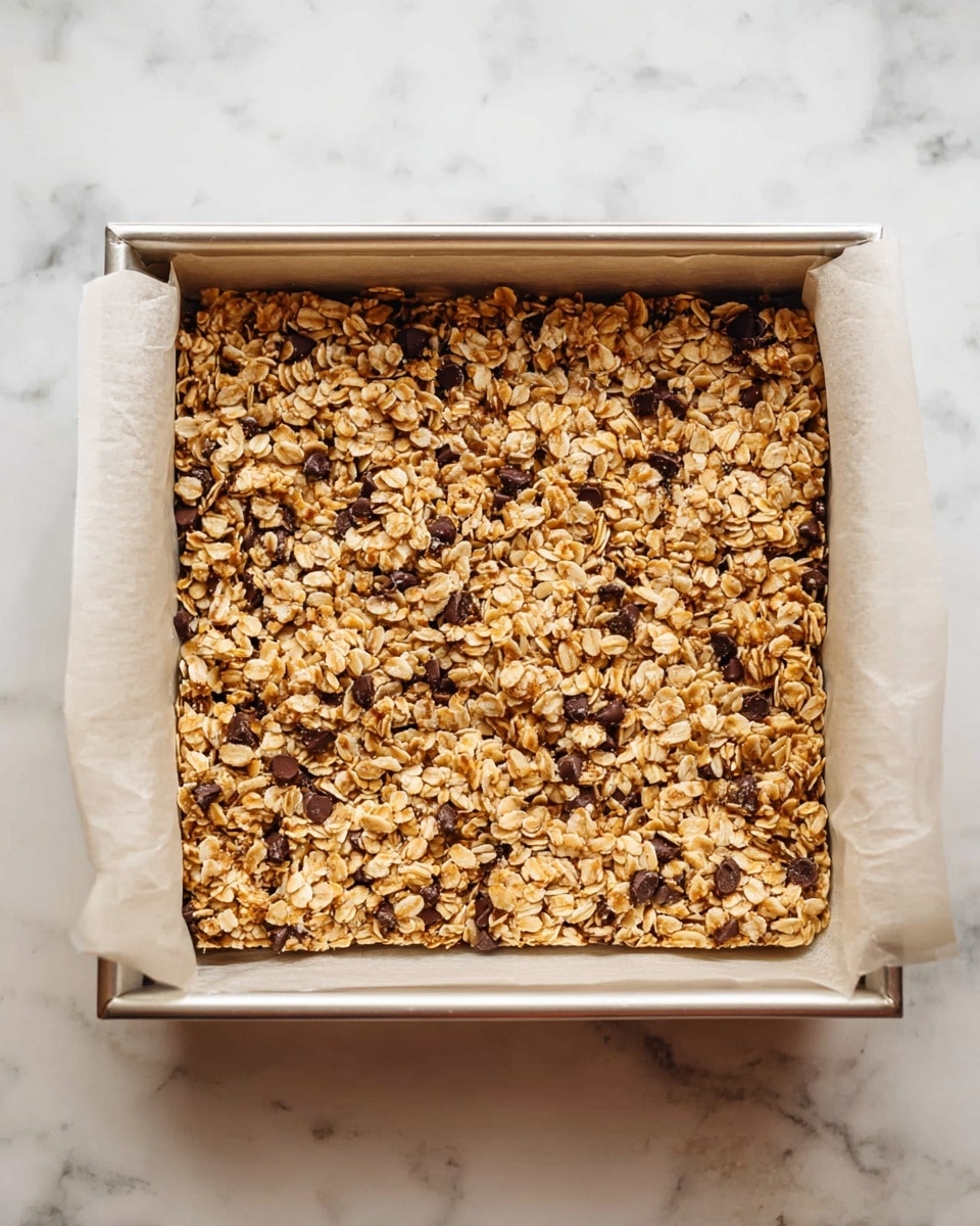 A square baking pan lined with parchment paper filled with one flat layer of golden brown granola bars mixed with visible dark chocolate chips, showing a rough and chunky texture from rolled oats and nuts evenly spread inside the pan. The pan rests on a white marbled surface. photo taken with an iphone --ar 4:5 --v 7