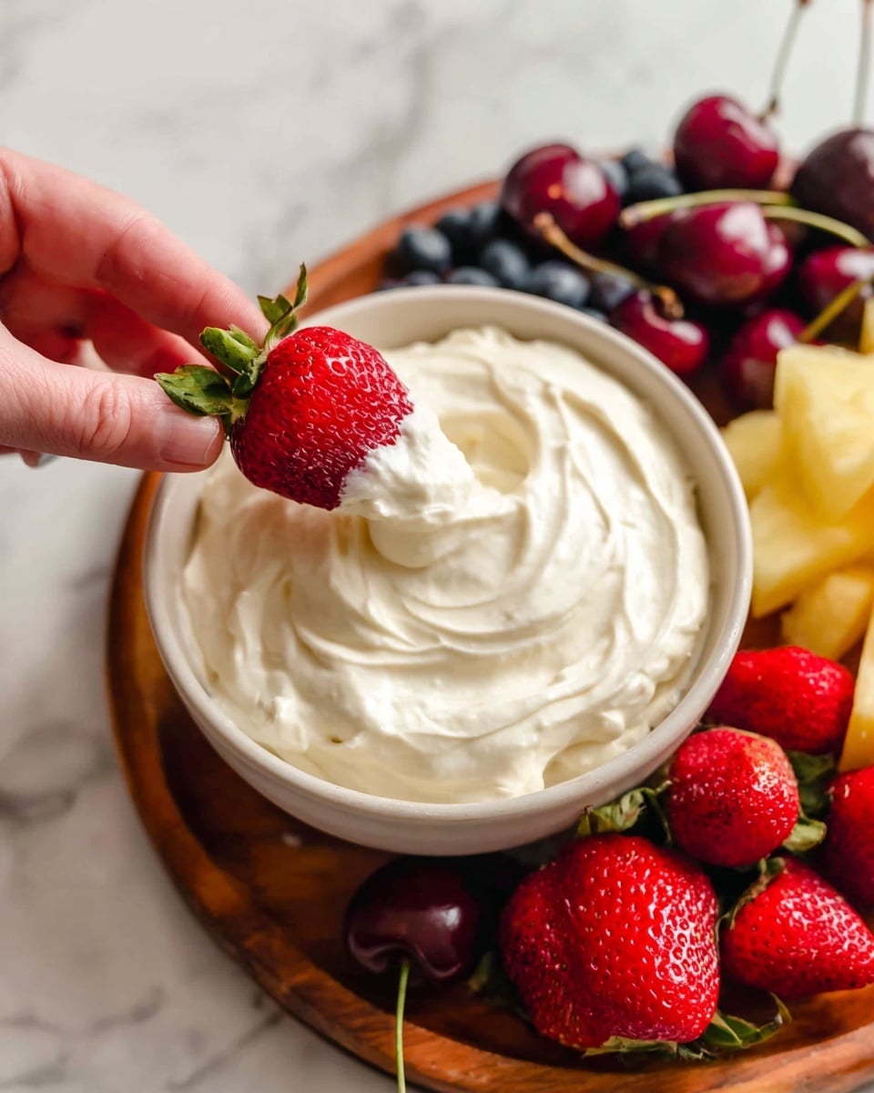 A close-up image shows a white bowl filled with smooth, creamy white dip with soft swirls on the surface, placed on a wooden board. Surrounding the bowl are fresh red strawberries, bright yellow pineapple chunks, dark red cherries with stems, and some blueberries partially visible at the top left. A woman's hand holds a bright red strawberry dipped halfway in the creamy white dip, showing the creamy texture clinging to the fruit against a white marbled surface background photo taken with an iphone --ar 4:5 --v 7