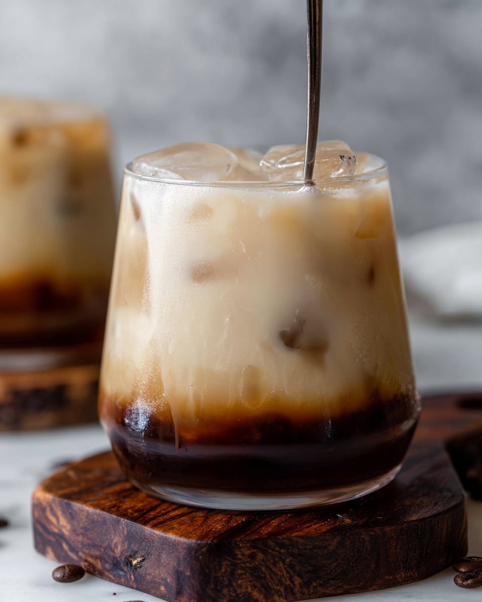 The image shows a close-up of a short, clear glass filled with iced coffee. The drink has three visible layers: a bottom dark brown layer from coffee, a middle creamy beige layer blending smoothly with the coffee, and a top light beige layer with ice cubes floating inside. A metal spoon stands vertically in the glass. The glass is placed on a dark wooden coaster set on a white marbled surface, with some coffee beans visible on the surface nearby. In the background, there is a blurry part of a similar glass. The photo taken with an iphone --ar 4:5 --v 7