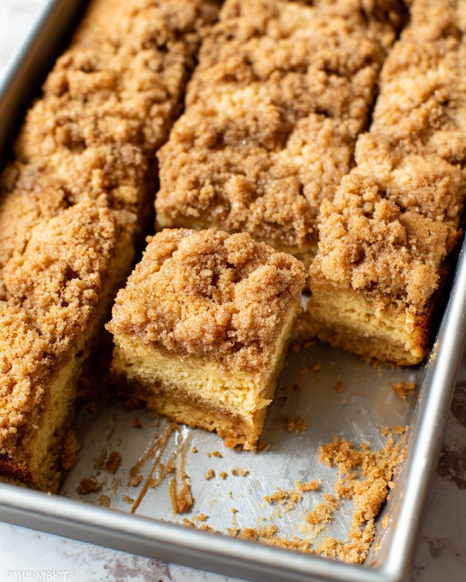 The image shows a baking pan with a crumb cake cut into squares. The cake has two visible layers: the base layer is a soft, golden brown cake with a smooth texture, while the top layer is a crumbly streusel topping with a light brown color and coarse texture. One piece is removed from the pan, revealing the cake's dense but tender inside. Crumbs are scattered inside the pan around the cut pieces. The baking pan has a silver metal finish, and the photo is taken from above with clear lighting, showing the texture of the crumb topping well. photo taken with an iphone --ar 4:5 --v 7