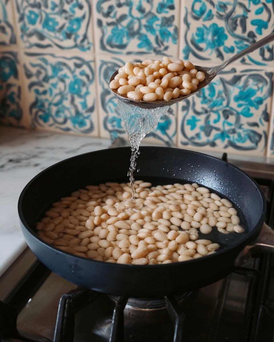 A close-up view of a black pan filled with soft light beige beans soaking in clear water, with a silver spoon above it holding more beans and some water dripping back into the pan. The pan is on a stove with black grates, and behind it is a wall with cream-colored tiles featuring turquoise blue and dark grey floral patterns. A white marbled surface is just visible at the bottom. Photo taken with an iphone --ar 4:5 --v 7