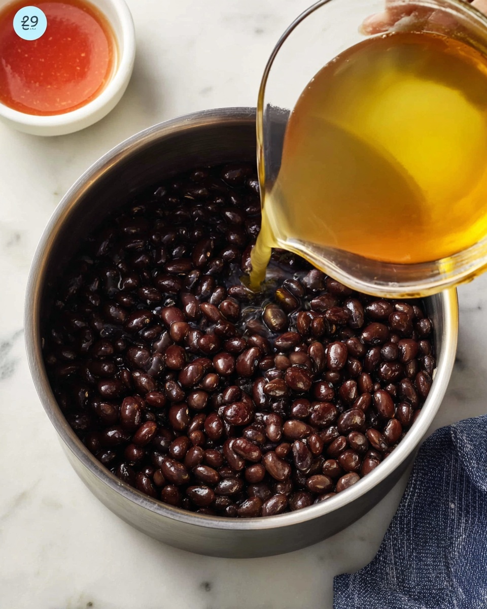 In this image, a stainless steel pot sits on a white marbled surface and is filled with a layer of dark brown black beans, shiny and cooked. A clear glass container, tilted above the pot, is pouring a light yellow broth or liquid onto the beans. In the top left corner, there is a small white bowl with a reddish sauce inside, partially visible. A blue and white kitchen towel is placed to the right side of the pot. The photo taken with an iphone --ar 4:5 --v 7