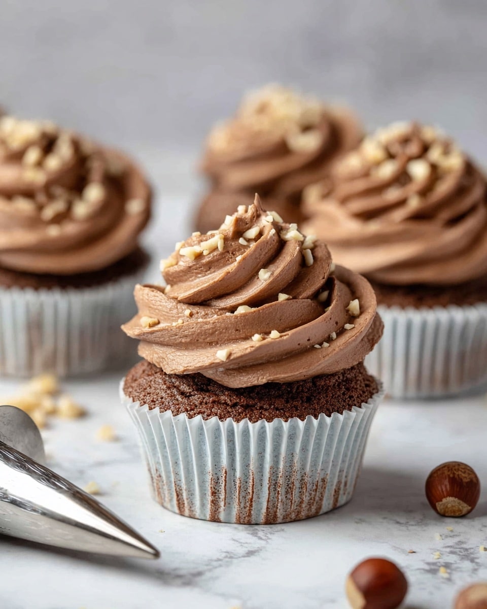The image shows four chocolate cupcakes on a white marbled surface, each topped with thick swirls of smooth light brown chocolate frosting arranged in two or three layers. The frosting is sprinkled with small chopped nuts that add texture and contrast. The cupcake liners are white with faint brown stripes, revealing the darker chocolate cake base beneath. In the foreground, a metal piping tip with some chocolate frosting around the edges lies next to a whole hazelnut and scattered nut pieces, adding a natural, fresh touch to the scene. The background is softly blurred, keeping focus on the closest cupcake and frosting details. Photo taken with an iphone --ar 4:5 --v 7
