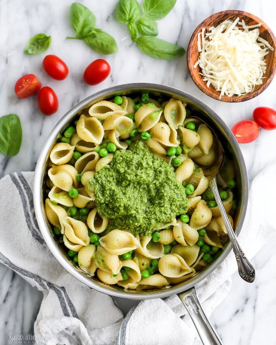 A shiny silver pot filled with pale yellow shell pasta mixed with bright green peas sits on a white marbled surface with a white and gray striped cloth underneath. In the center of the pasta, there is a thick, green pesto sauce layer covering some pasta shells. Around the pot, there are a few scattered cherry tomatoes and green basil leaves, with a small wooden bowl filled with grated cheese placed nearby. A silver spoon rests inside the pot on the right side. The whole scene is bright and fresh, emphasizing the colors and textures of the pasta dish. photo taken with an iphone --ar 4:5 --v 7
