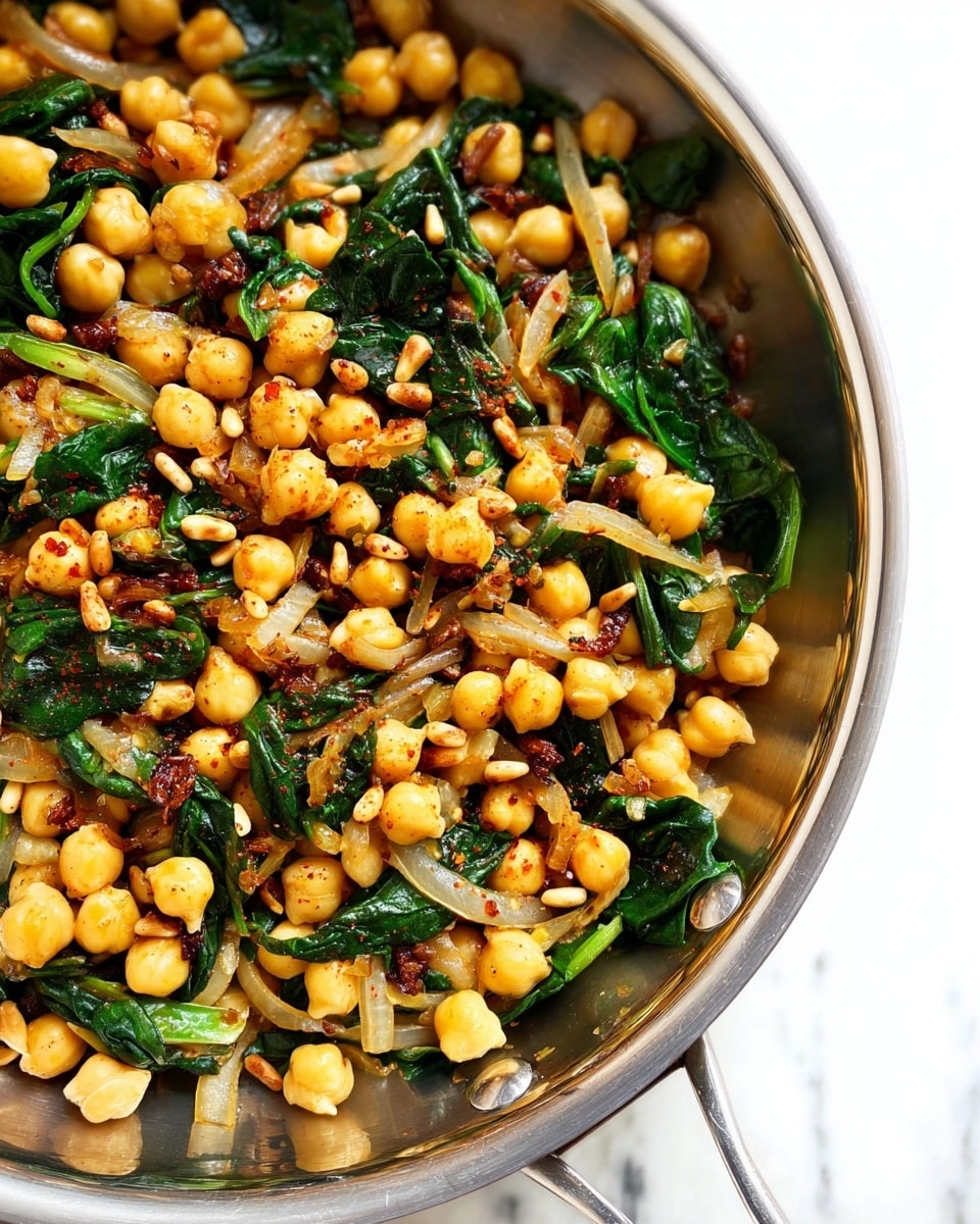 The image shows a close-up view of a stainless steel pan filled with a colorful chickpea dish. The chickpeas are light yellow and mixed with dark green wilted spinach leaves and thin, translucent onion slices. Small brown raisins and light tan pine nuts are spread throughout, adding texture and contrast. A sprinkle of red spice powder gives a subtle reddish tint on some chickpeas and onions. The pan’s shiny metal surface reflects light, with part of the handle visible on the left side. The background is a white marbled texture. photo taken with an iphone --ar 4:5 --v 7