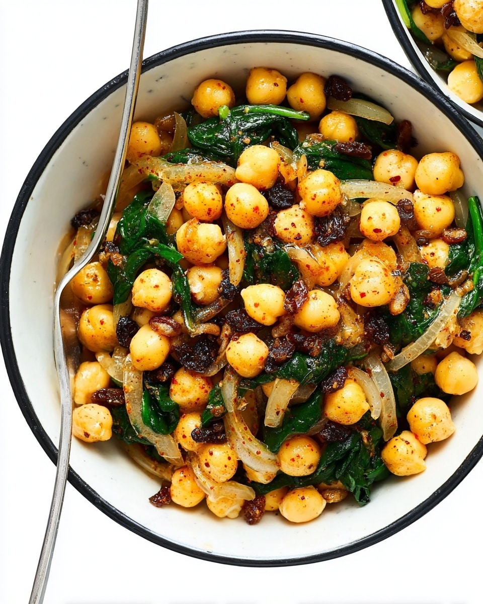 The image shows a white bowl with a black rim filled with a colorful chickpea salad. The base layer is light yellow chickpeas, mixed with wilted dark green spinach leaves and translucent cooked onion slices. Scattered over and mixed within are small pale pine nuts and dark brown raisins, all lightly coated with a reddish seasoning and black pepper. A silver spoon rests inside the bowl on the left side. The background surface is white with a light marbled texture. Photo taken with an iphone --ar 4:5 --v 7
