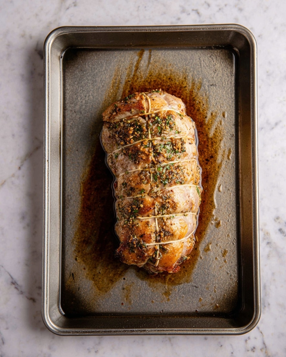 A single piece of roasted meat sits in the center of a rectangular metal baking tray. The meat is tied with string in a segmented pattern, showing a rounded, plump shape with a light golden-brown color and a surface sprinkled with green herbs and small bits of seasoning. The tray shows streaks of dark brown cooking juices and oil residue around the meat. The scene is set on a white marbled surface. photo taken with an iphone --ar 4:5 --v 7