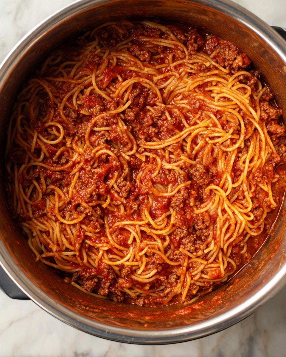 The dish shows a large pot filled with spaghetti mixed with a rich red tomato sauce that covers most of the noodles. There are small pieces of ground meat spread evenly throughout the pasta, giving it a textured look with bits of darker brown meat contrasting with the red sauce and light orange noodles. The spaghetti strands are long, thin, and tangled together, soaking in the sauce, with some parts slightly shiny from the moisture. The inside of the pot is metallic, and the photo is taken on a white marbled surface. photo taken with an iphone --ar 4:5 --v 7