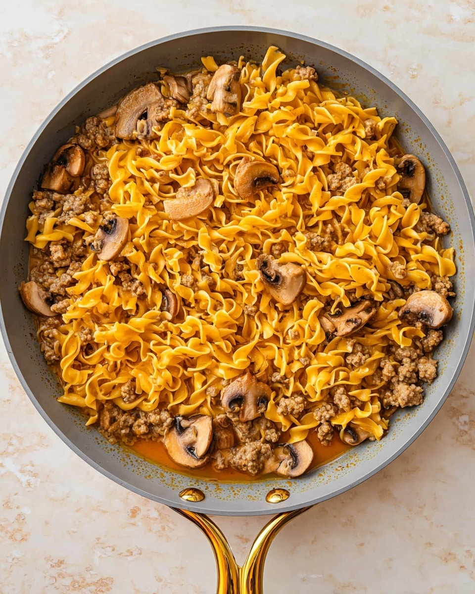 A gray pan with a golden handle holds a one-layer dish of bright yellow curly egg noodles mixed with light brown cooked ground meat and medium brown sliced mushrooms. The ingredients are spread evenly throughout the pan, with some noodles sitting in a thin, orange sauce that fills the pan partially. The pan rests on a white marbled surface. photo taken with an iphone --ar 4:5 --v 7