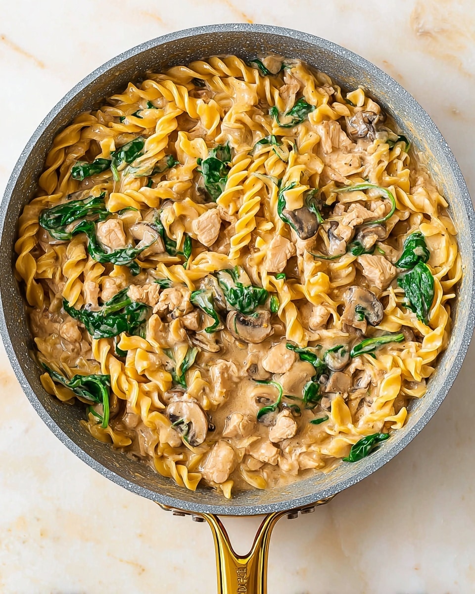 The image shows a pan filled with a creamy pasta dish. The base layer consists of yellow twisted egg noodles mixed evenly with small pieces of light brown cooked turkey and chunks of soft, thickly sliced tan mushrooms. Bright green spinach leaves are scattered throughout, adding a fresh color contrast. The creamy sauce has a light beige tone, coating the noodles and ingredients smoothly. The pan has a gray interior with a shiny gold handle, sitting on a white marbled surface. photo taken with an iphone --ar 4:5 --v 7