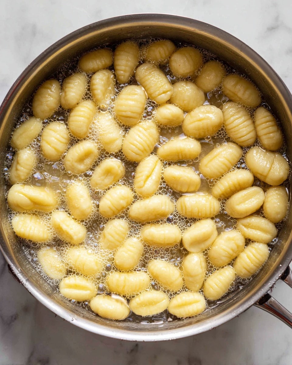 The image shows a silver pan filled with many small, oval-shaped gnocchi pieces cooking in bubbling water or butter. The gnocchi are light yellow with a soft, smooth texture and have ridged or slightly curved patterns on their tops. The pan sits on a white marbled surface, and the scene focuses closely on the gnocchi inside the pan, showing their plump, tender form and the shiny, hot liquid around them. photo taken with an iphone --ar 4:5 --v 7