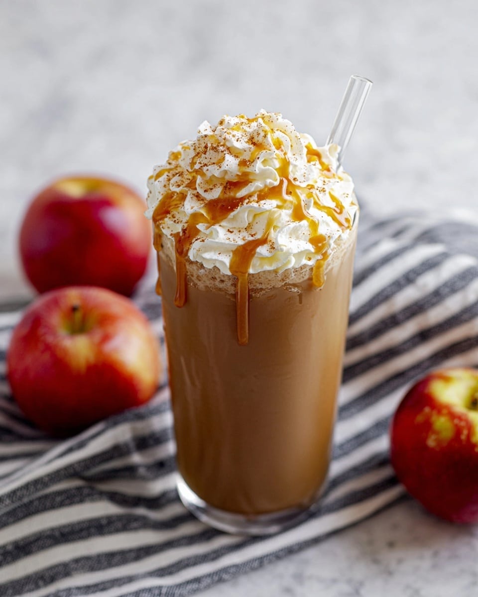 A tall clear glass filled with a smooth light brown drink topped with a large swirl of white whipped cream. The whipped cream is drizzled with golden caramel sauce and sprinkled with a light dusting of cinnamon. A clear glass straw is placed on the right side of the glass, leaning outward. The glass rests on a white marbled surface, with two red apples and a black and white striped cloth behind it. Photo taken with an iphone --ar 4:5 --v 7