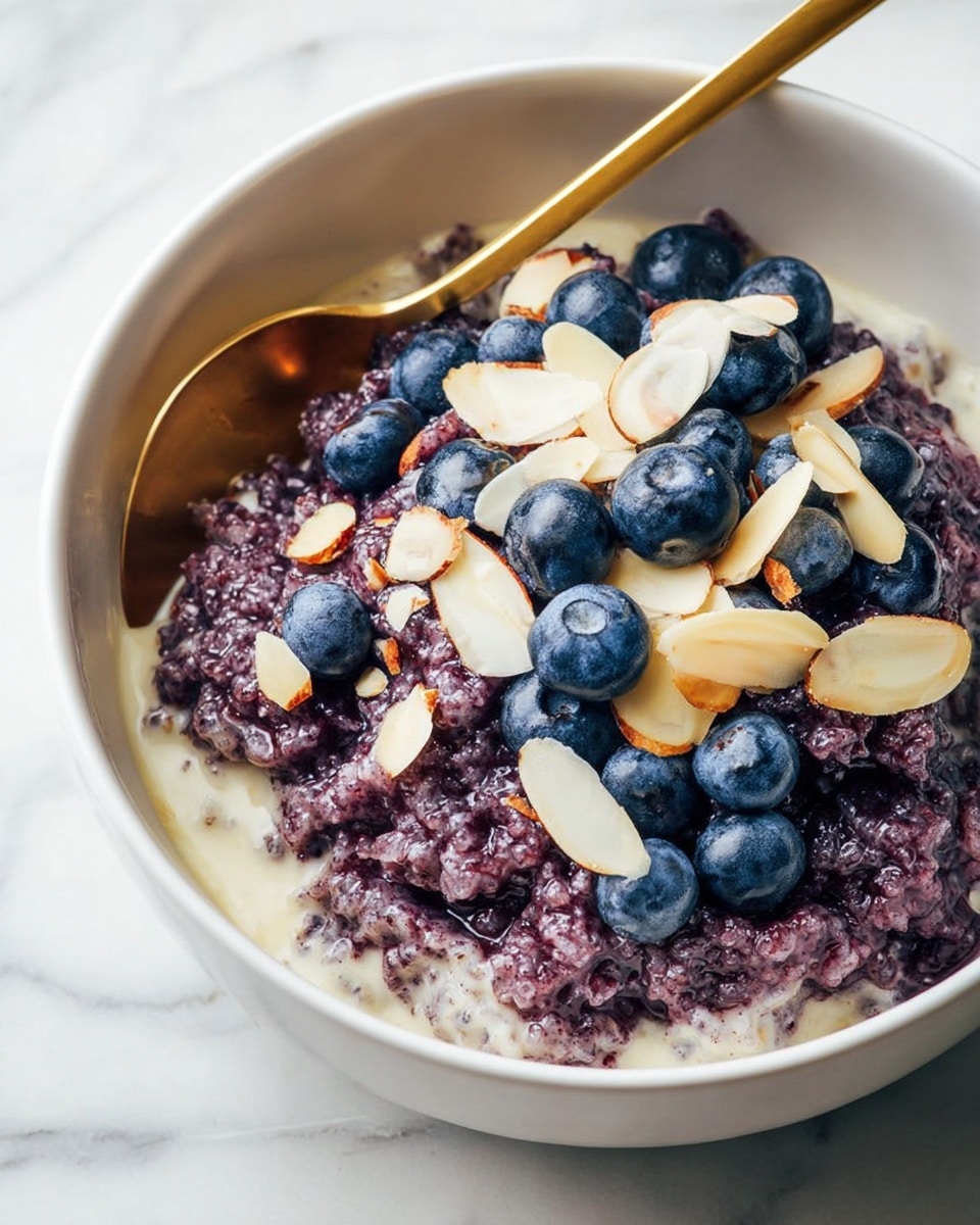 A white bowl filled with creamy light beige milk at the bottom, topped with a thick layer of dark purple rice pudding with a grainy and slightly wet texture. Scattered on top are fresh, plump blueberries in deep blue color and thin, pale almond slices. A shiny golden spoon rests inside the bowl, partly submerged in the pudding, adding a bright contrast. The bowl sits on a white marbled surface. Photo taken with an iphone --ar 4:5 --v 7
