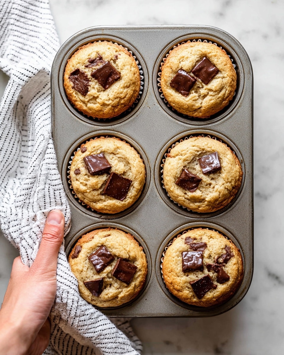 A metal muffin tray with six freshly baked muffins inside, each muffin topped with three to five large dark chocolate chunks that are slightly melted and shiny. The muffins have a light golden-brown color with a soft, slightly cracked surface showing a tender texture. They fill the muffin cups fully, with edges slightly darker and firm. The tray sits on a white marbled surface. In the lower-left corner, a woman's hand holds a white cloth with thin black stripes, gripping the tray from the side. Photo taken with an iphone --ar 4:5 --v 7